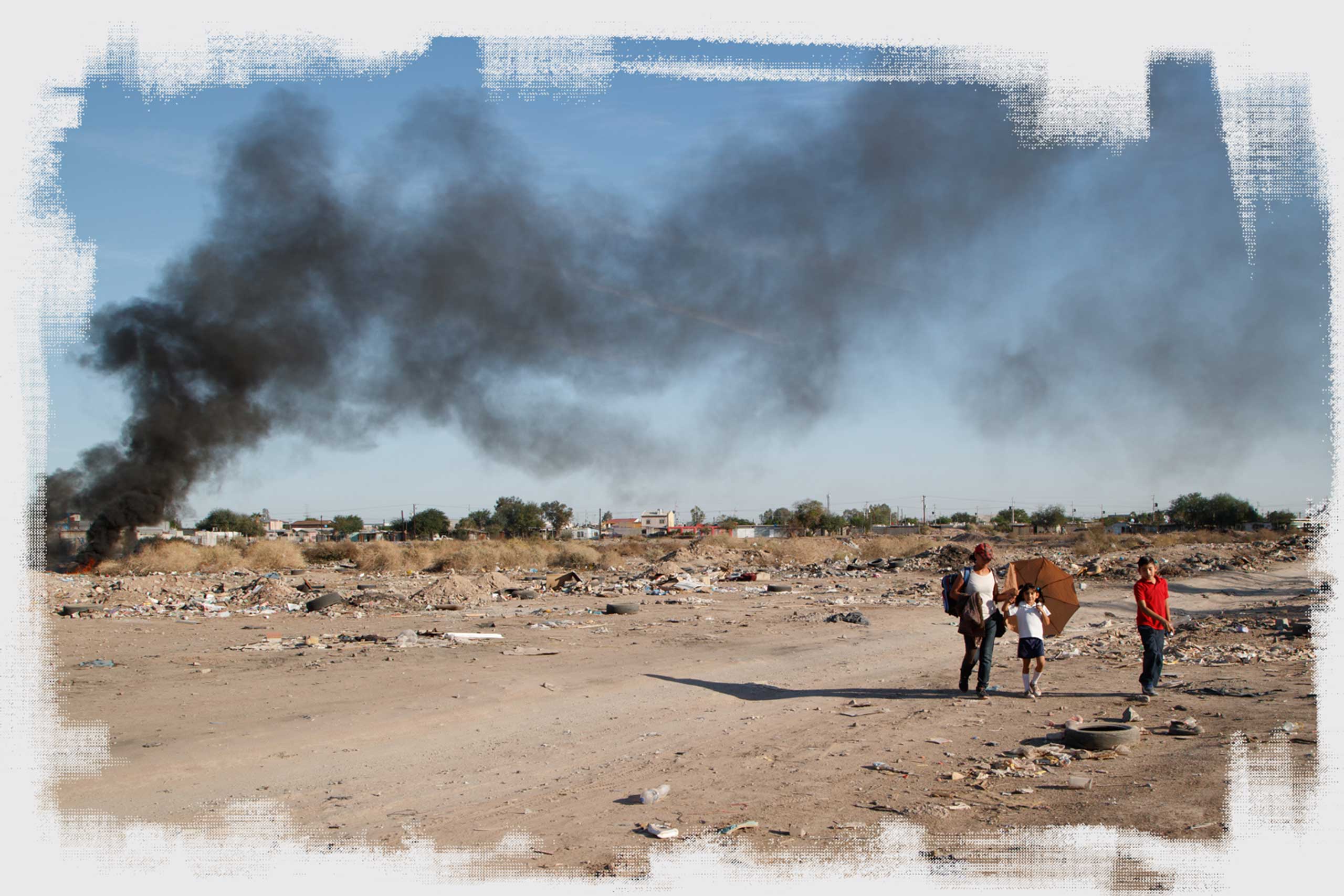 A woman walks two children home from school through a vacant lot in Mexicali, passing a heap of burning tires and trash.