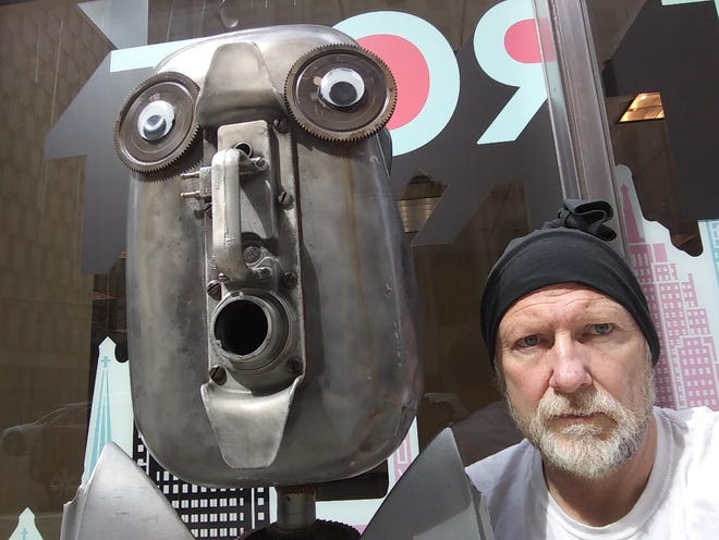 Tim Burke poses with one of his scrap-and-spare-parts robots, installed on Fort Street  near Griswold in downtown Detroit.