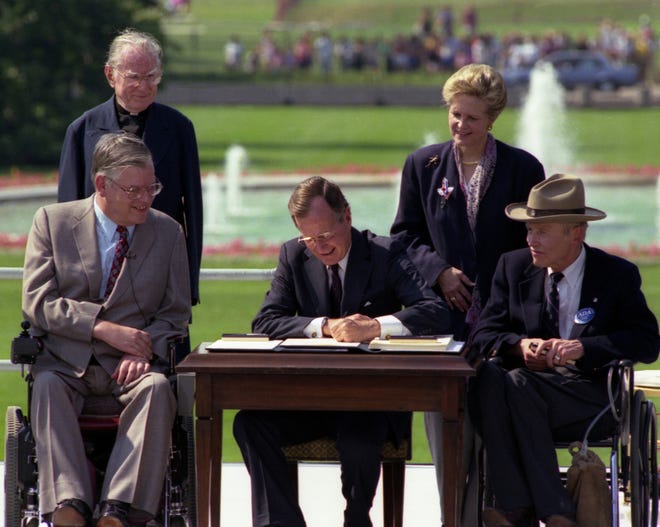 President Bush signs the Americans with Disabilities Act on the South Lawn of the White House on July 26, 1990. With him are the Rev. Harold Wilkie of California, standing at left; Sandra Parrino of the National Council on Disability, standing right; Evan Kemp, Chairman of Equal Opportunity Commission, seated left; and Justin Dart of the Presidential Commission on Employment of People with Disabilities.