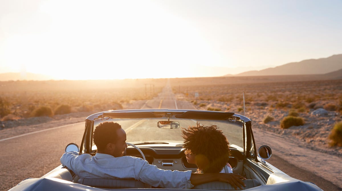 Rear View Of Couple On Road Trip Driving Classic Convertible Car Towards Sunset