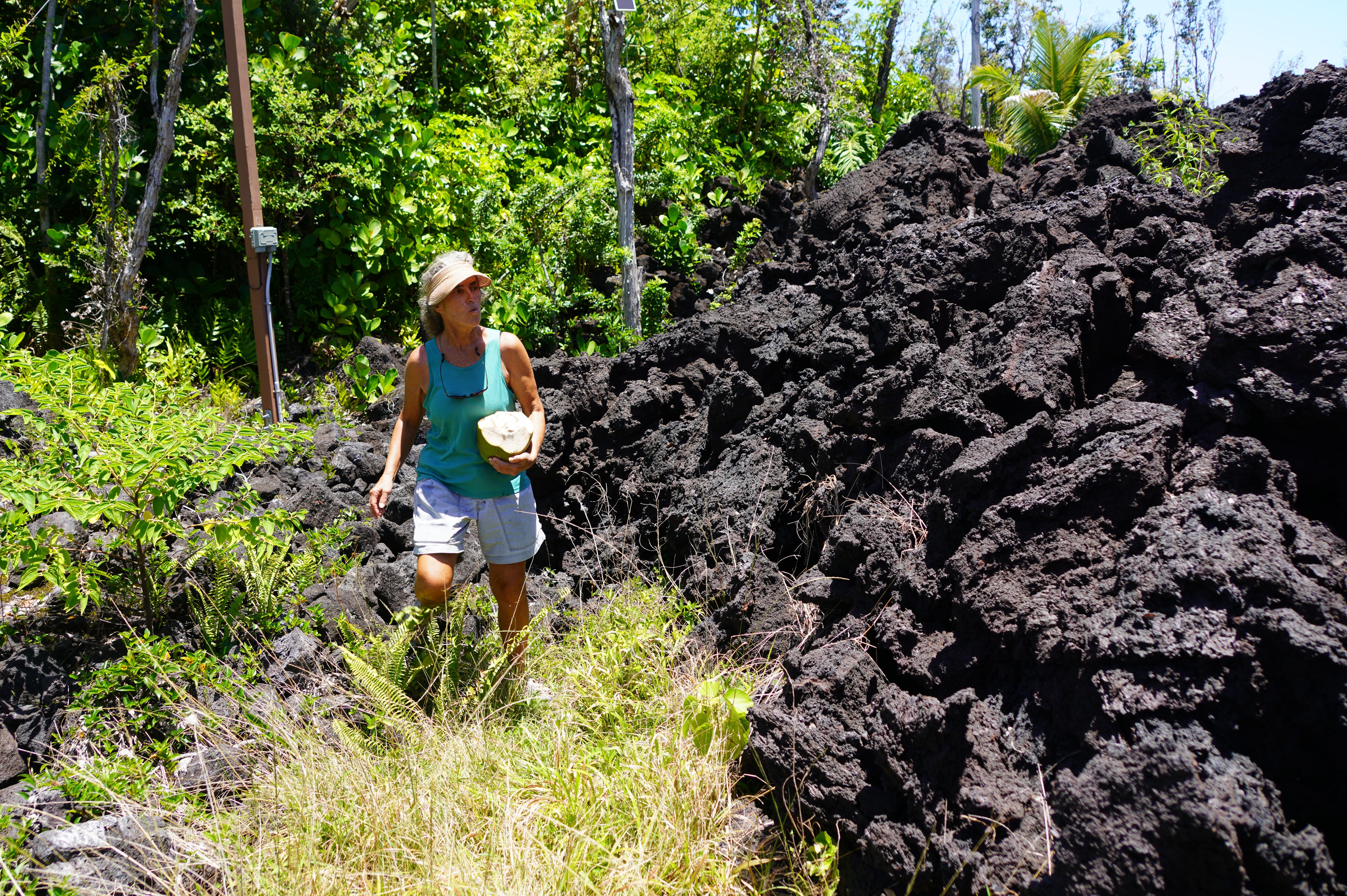 Deb Smith, holding a freshly opened coconut, searches for the front gate of her fruit farm on Hawaii's Big Island. The Smith's property was covered with about 50 feet of lava during the 2018 eruption.