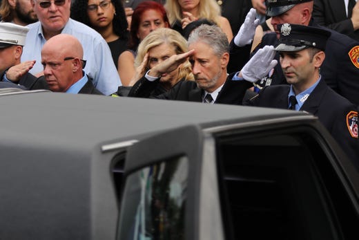 Comedian and activist Jon Stewart (C) and U.S. Rep. Carolyn Maloney (D-NY) (C-L) salute the casket of retired NYPD detective Luis Alvarez as it is brought into Immaculate Conception Church on July 3, 2019 in the Queens borough of New York City. Alvarez, who passed away Saturday at the age of 53 following a three-year battle with colorectal cancer, became ill following the months he spent exposed to toxins at the Ground Zero site after the Twin Towers collapsed. He became a national figure after he spent his last days fighting for the September 11th Victim Compensation Fund which pays for health benefits for the police, firefighters and others who have become sick after being exposed to toxins following the September 11 attacks.