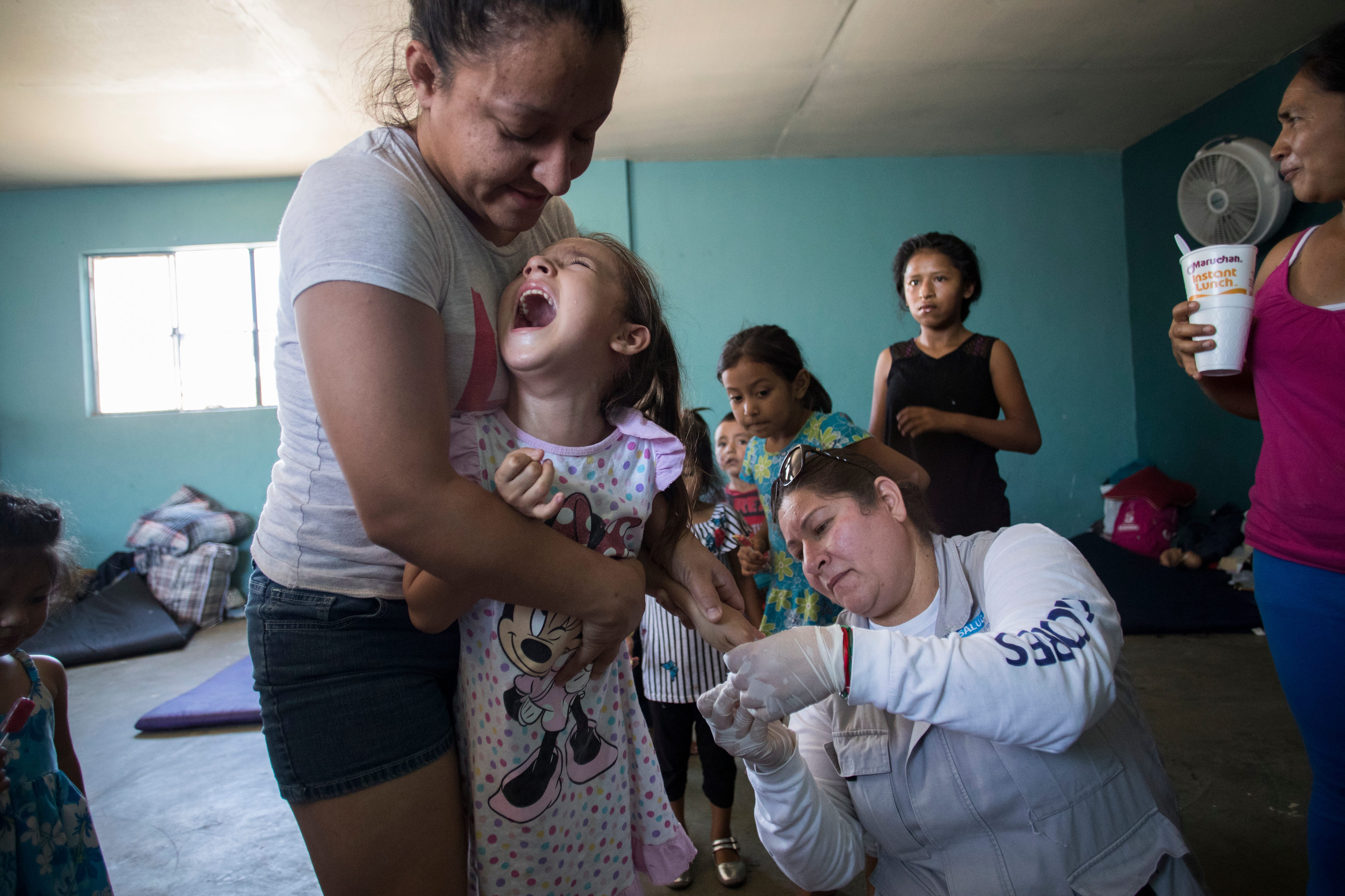 MEXICALI, Mexico – Marisela Lopez, 28, holds her 6-year-old daughter, Maritza Salazar Lopez, as Andrea Delgado with the Baja California state health ministry draws blood from her finger on June 24, 2019. Delgado was testing for malaria at the migrant shelter Casa de Ayuda Alfa y Omega.