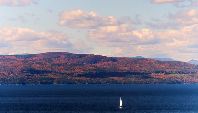 The white sails of a sloop on Lake Champlain stands in contrast to the Adirondacks bathed in fall colors seen from the Vermont side.