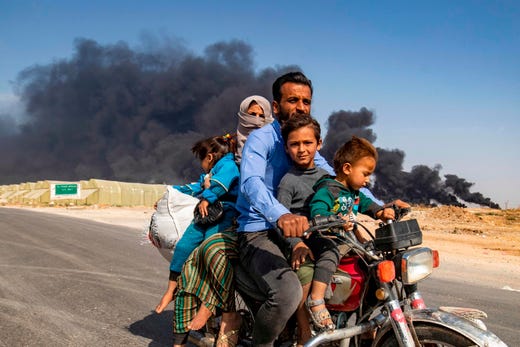 TOPSHOT - Displaced people, fleeing from the countryside of the Syrian Kurdish town of Ras al-Ain along the border with Turkey, ride a motorcycle together along a road on the outskirts of the nearby town of Tal Tamr on October 16, 2019 as they flee from the Turkish 'Peace Spring' military operation, with smoke plumes of tire fires billowing in the background to decrease visibility for Turkish warplanes in the area. (Photo by Delil SOULEIMAN / AFP) (Photo by DELIL SOULEIMAN/AFP via Getty Images) ORIG FILE ID: AFP_1LH14T