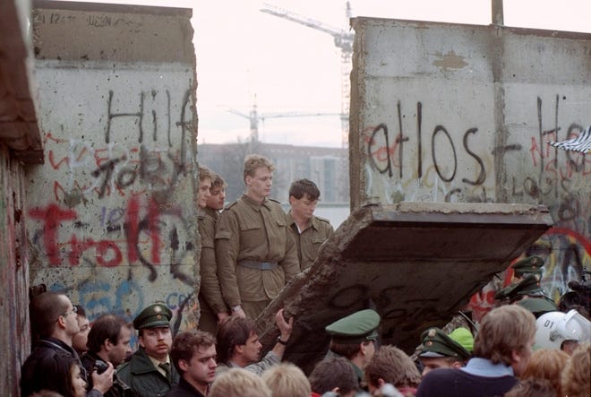 East German border guards look through a hole in the Berlin wall after demonstrators pulled down one segment of the wall at Brandenburg gate Saturday, November 11, 1989.