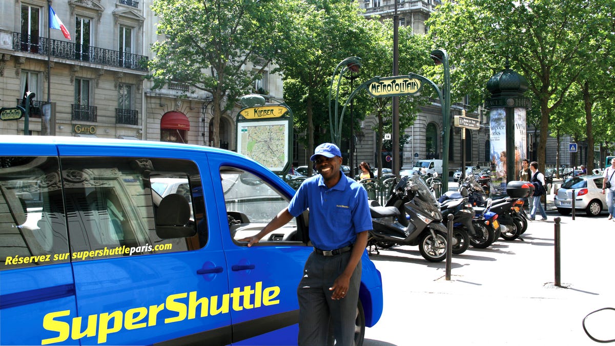 Airport Checkin -- SuperShuttle, a shared airport shuttle service, launched its first international service in Paris. Pictured is a SuperShuttle vehicle and its driver, Elie Tambou in Paris. Its service is available from Charles de Gaulle, Paris Orly and the local airport Beauvais to anywhere in Paris, including hotels and local residences.  (Via MerlinFTP Drop)