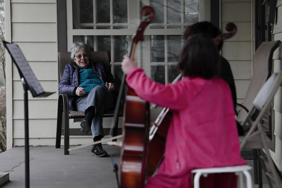 Helena Schlam, left, listens as her neighbors, Taran Tien, second from right, and Calliope Tien, right, play cello on her front porch on Monday, March 16, 2020, in Columbus, Ohio. The siblings decided to play a concert for Schlam because she was self-isolating due to the ongoing COVID-19 pandemic.