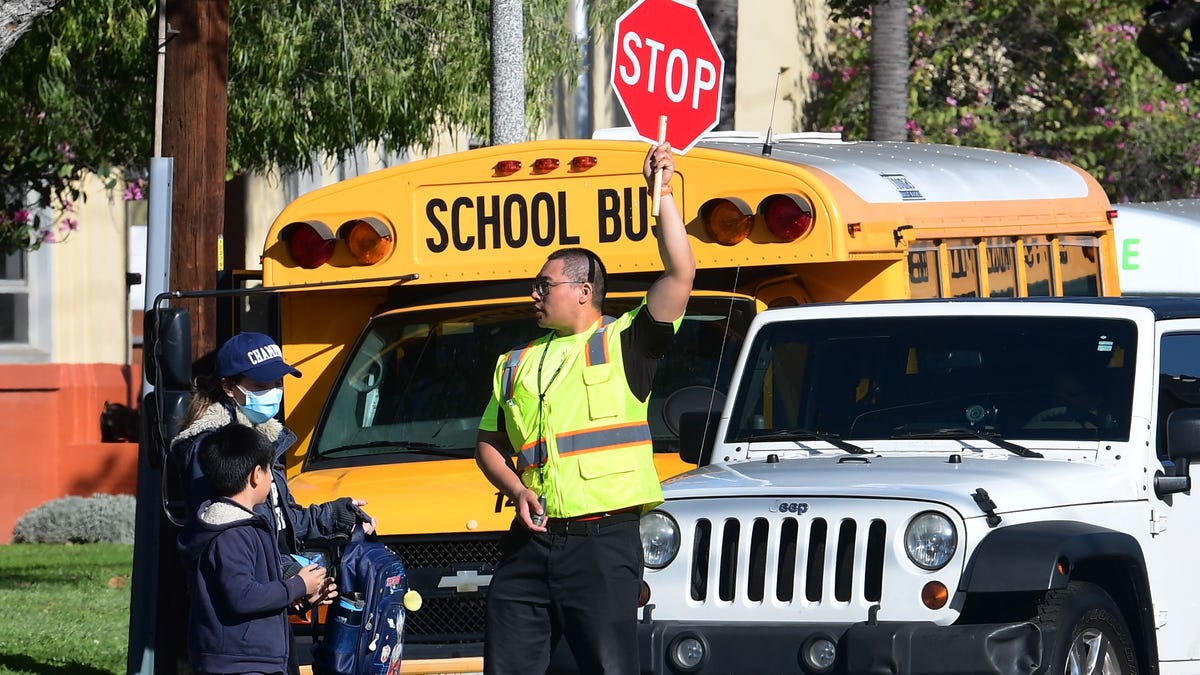 A woman wears a facemask picking up a child as an Alhambra Unified School District crossing guard stops traffic outside Ramona Elementary School on February 4, 2020 in Alhambra, California. - As the coronavirus outbreak  spreads, fuelling rumors and misinformation, a petition to cancel all classes in one US school district for fear of the virus has garnered nearly 14,000 signatures. The online petition posted on Change.org urges the Alhambra Unified School District   located east of Los Angeles and with a heavily Asian population, to basically shut down until the outbreak is over. School district officials, however, have dismissed the petition as a bid to whip up hysteria over the deadly outbreak that has killed hundreds in China.