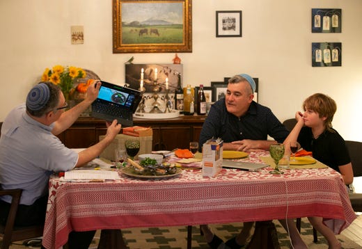 Rabbi Dean Shapiro (left) of Temple Emanuel in Tempe, angles his laptop so others online can see their Seder plate as Shapiro's partner, Haim Ainsworth and their son, Jacob Shapiro-Ainsworth, 11, look on, as they participate in an online Seder during the first night of the Jewish holiday of Passover at their home in Tempe on April 8, 2020. The Seder which included members from Temple Emanuel was being held online because of the coronavirus pandemic.