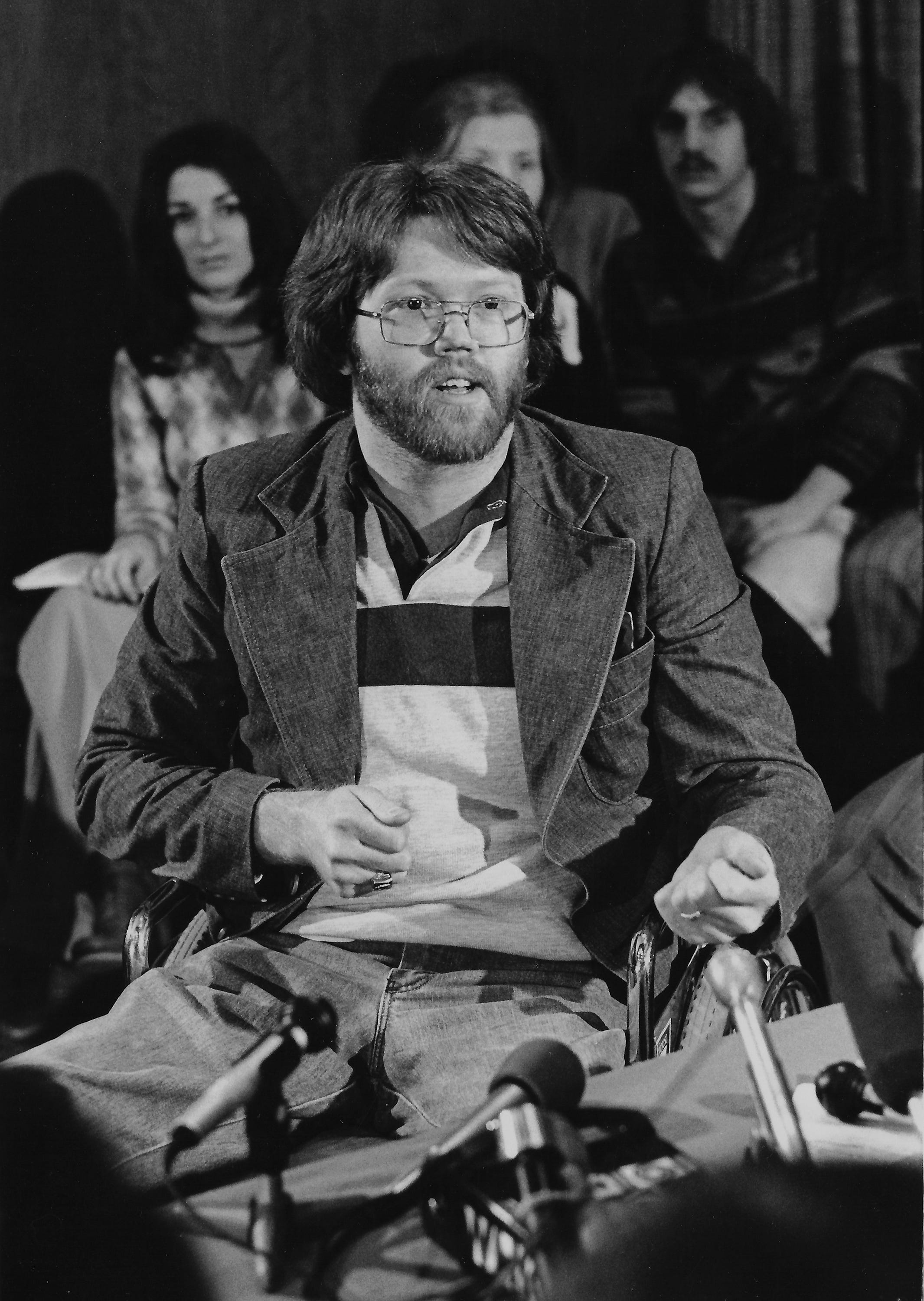 Dean Kahler, who was shot in the back and paralyzed by National Guardsmen at Kent State on May 4, 1970, speaks at a press conference following the $675,000 settlement of a civil lawsuit on Jan. 4, 1979. In the background at left is Roseanne “Chic” Canfora, who was a witness to the shootings and whose brother, Alan, was one of the nine students injured that day. [Ott Gangl/AKRON BEACON JOURNAL]