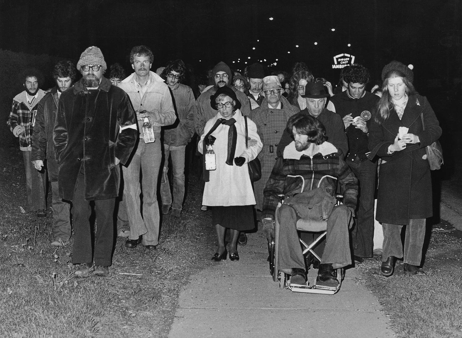 Professor Jerry Lewis (left, with beard), Elaine Holstein, mother of slain student Jeffrey Miller (center in white coat), and survivor Dean Kahler (in wheelchair) participate May 3, 1976, in the annual candlelight vigil march to commemorate the 1970 shootings on the Kent State campus.