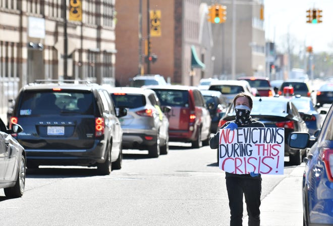 Organizer Joe McGuire walks and holds a sign while people drive their vehicles around Cadillac Place as part of a caravan protest organized by Detroit Eviction Defense in Detroit on May 13, 2020. The group calls for extending the eviction ban until at least 60 days have passed after the end of the current state of emergency.