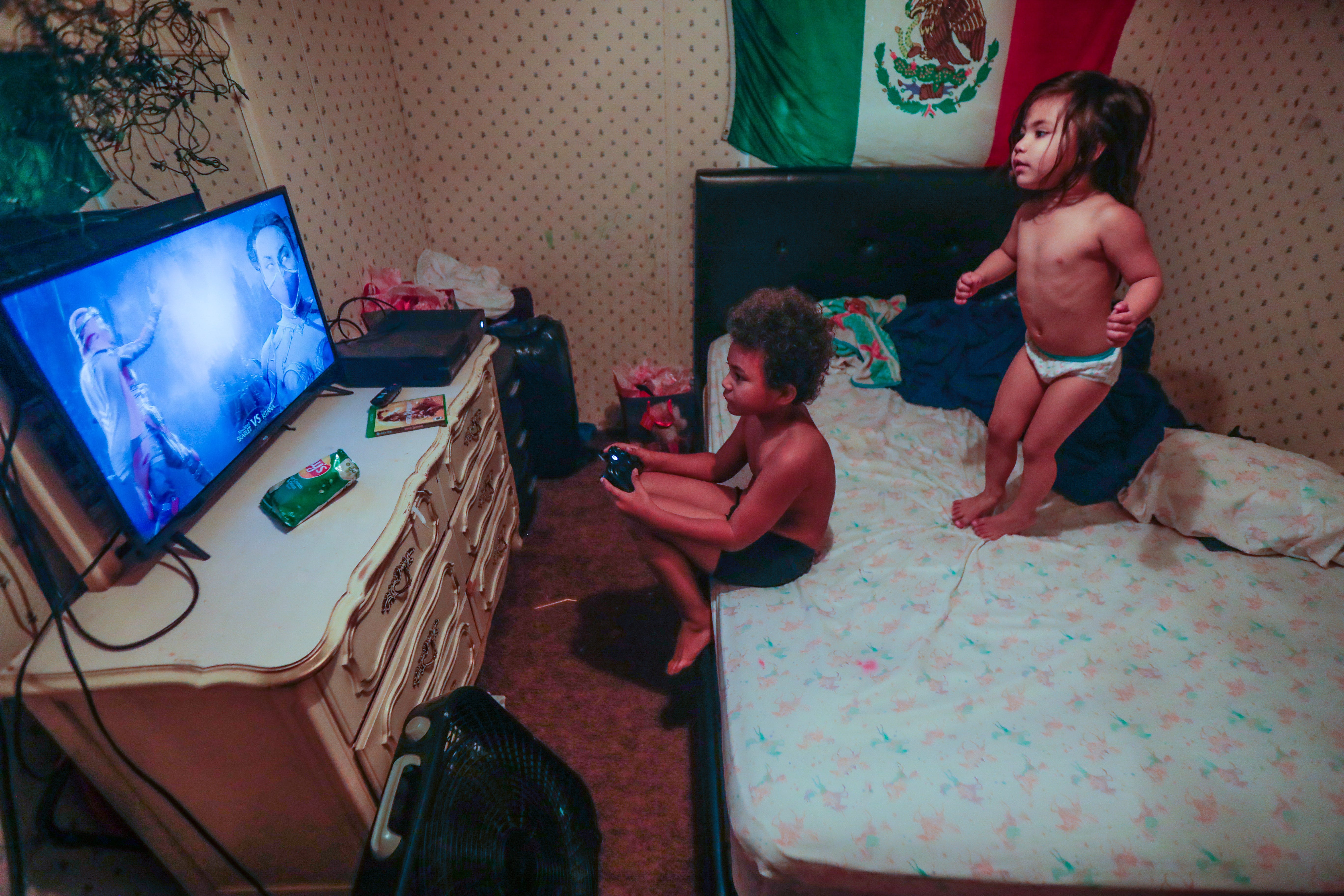 Victoria, 3, jumps on her mom's bed while her brother Romeo, 8, plays video games on his Xbox. The siblings live with their mom, her younger sister, and their mom's husband.