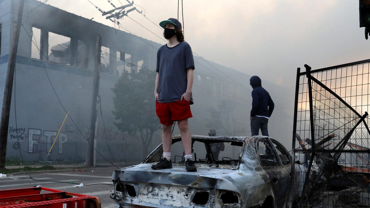 People stand on a burned up car as fires burn near a Target Store after a night of unrest and protests in the death of George Floyd early Thursday, May 28, 2020 in downtown Minneapolis.  Floyd died after being restrained by Minneapolis police officers on Memorial Day. A video taken by a bystander shows a Minneapolis police officer with his knee on the neck of a man in custody who later died. The four officers involved have been fired.