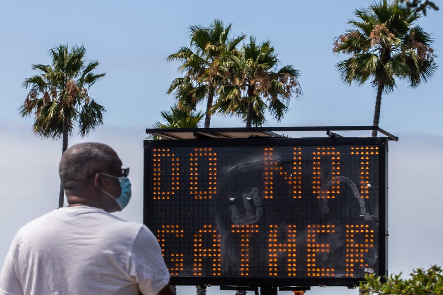 A man wearing a facemask checks his phone near a sign urging people not to gather, while he walks on the beach in Long Beach, Calif. on July 14, 2020.  California's Governor Gavin Newsom announced a significant rollback of the state's reopening plan on July 13, 2020 as coronavirus cases soared across America's richest and most populous state.