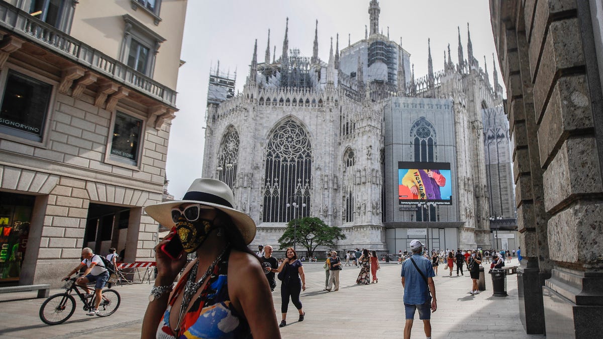 Pedestrians pass by a screen on the Duomo cathedral, showing a Moschino model during the Milan Digital Fashion Week, in Milan, Italy, Tuesday, July 14, 2020. Forty fashion houses are presenting previews of menswear looks for next spring and summer and pre-collections for women in digital formats, due to concerns generated by the COVID-19.