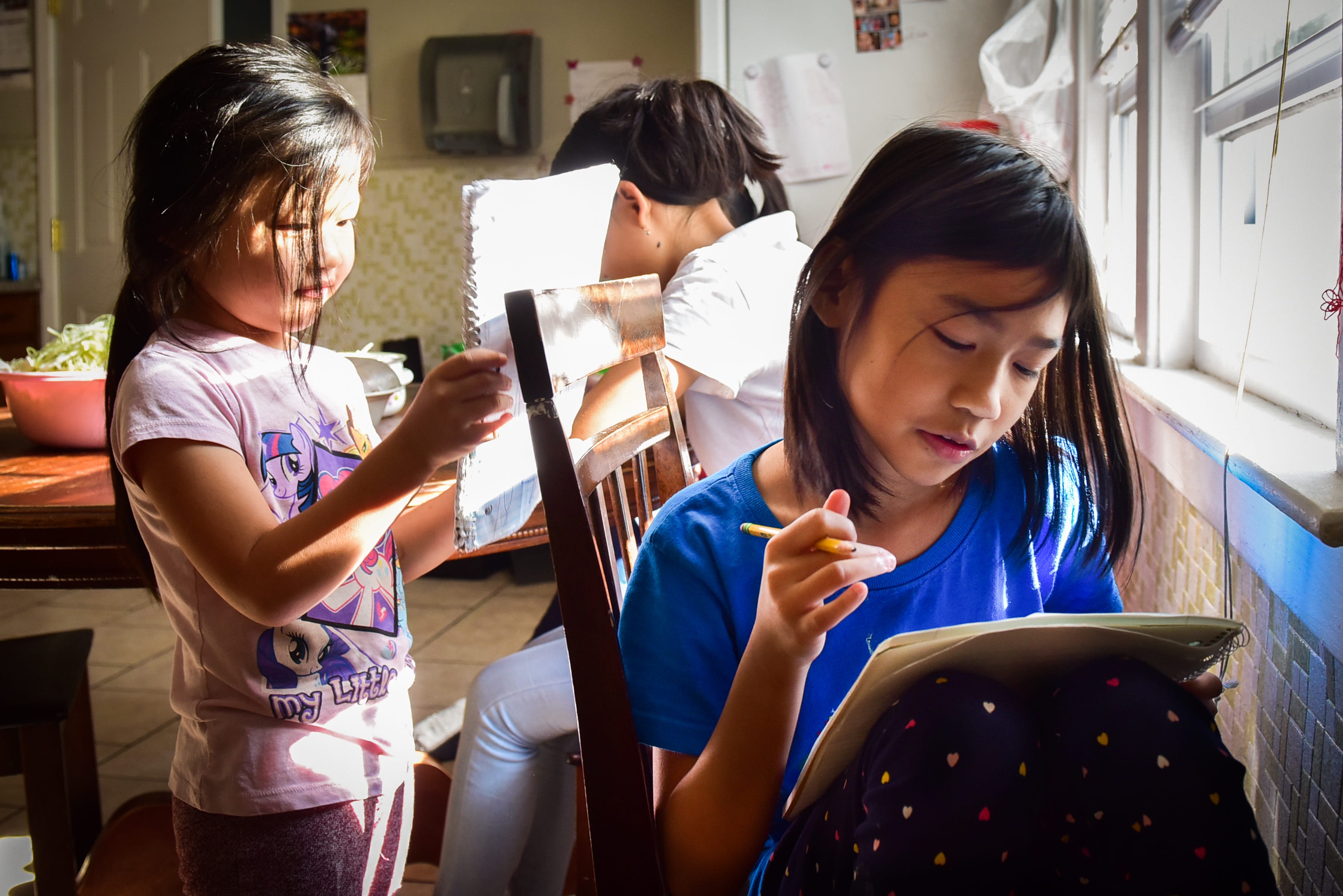 Eh Moo Taw Heh, right, draws in a notebook while her family prepares dinner in the kitchen on Sunday, Oct. 25, 2020 at their home on Utica. Eh Moo says her favorite class to take while learning from home is art.