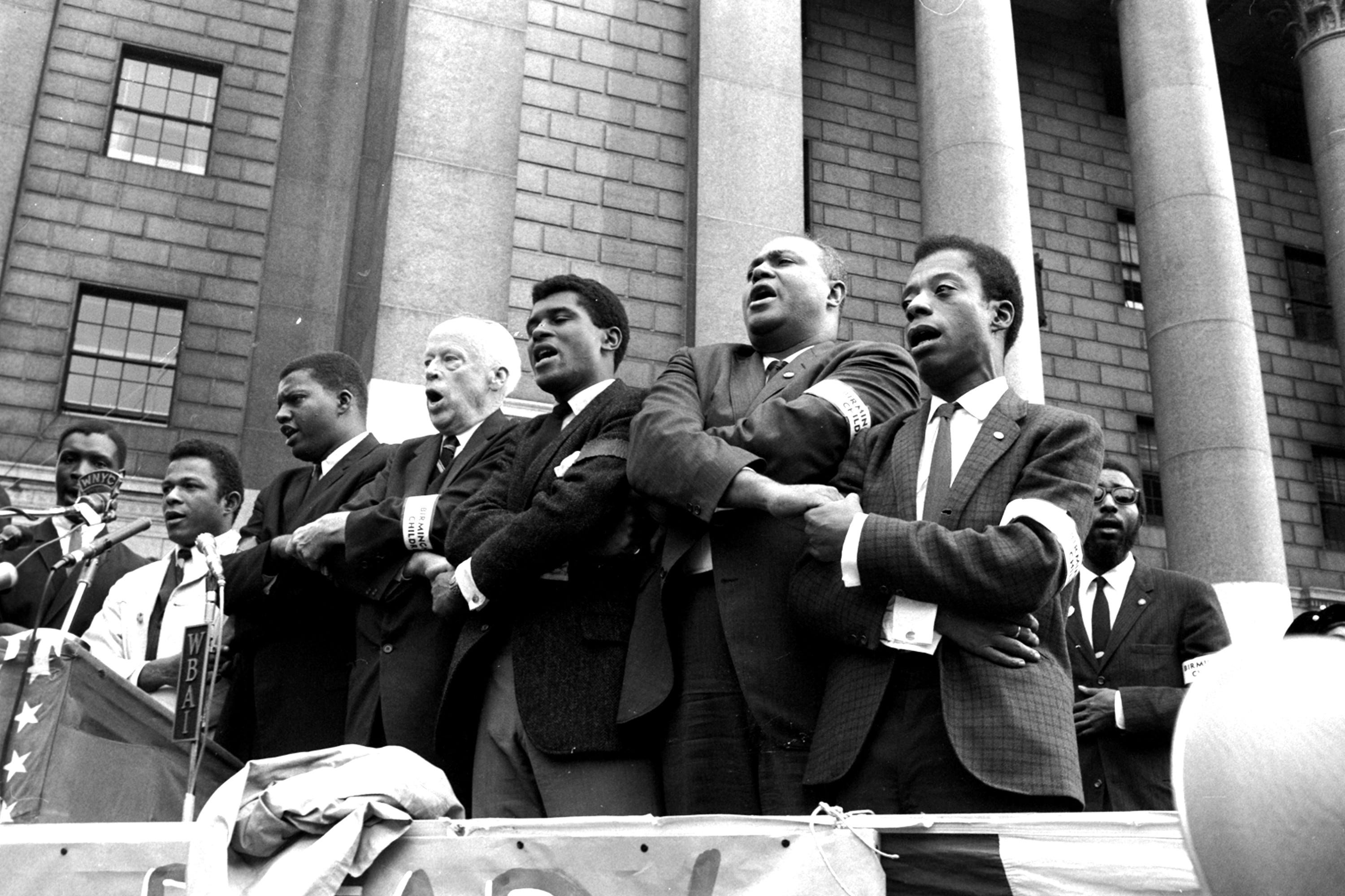 Civil rights leaders clasp hands as they sing during the closing of memorial ceremonies in New York, Sept. 22, 1963, for four girls killed in the bombing of the Birmingham, Ala., Sixteenth Street Baptist Church.  From right, are: author James Baldwin; James Farmer, president of the Congress of Racial Equality; unidentified man; and veteran Socialist leader Norman Thomas.  At a rally on Foley Square, Farmer charged the U.S. Department of Justice and President Kennedy with a share of the blame in the Sept. 15, bombing, and Thomas said he was "ashamed that I was a white American,  when the bombing took place."