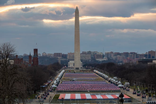 Against a city skyline and cloudy sky, a field of red and blue flags is arranged in many rows before a large white pillar. 