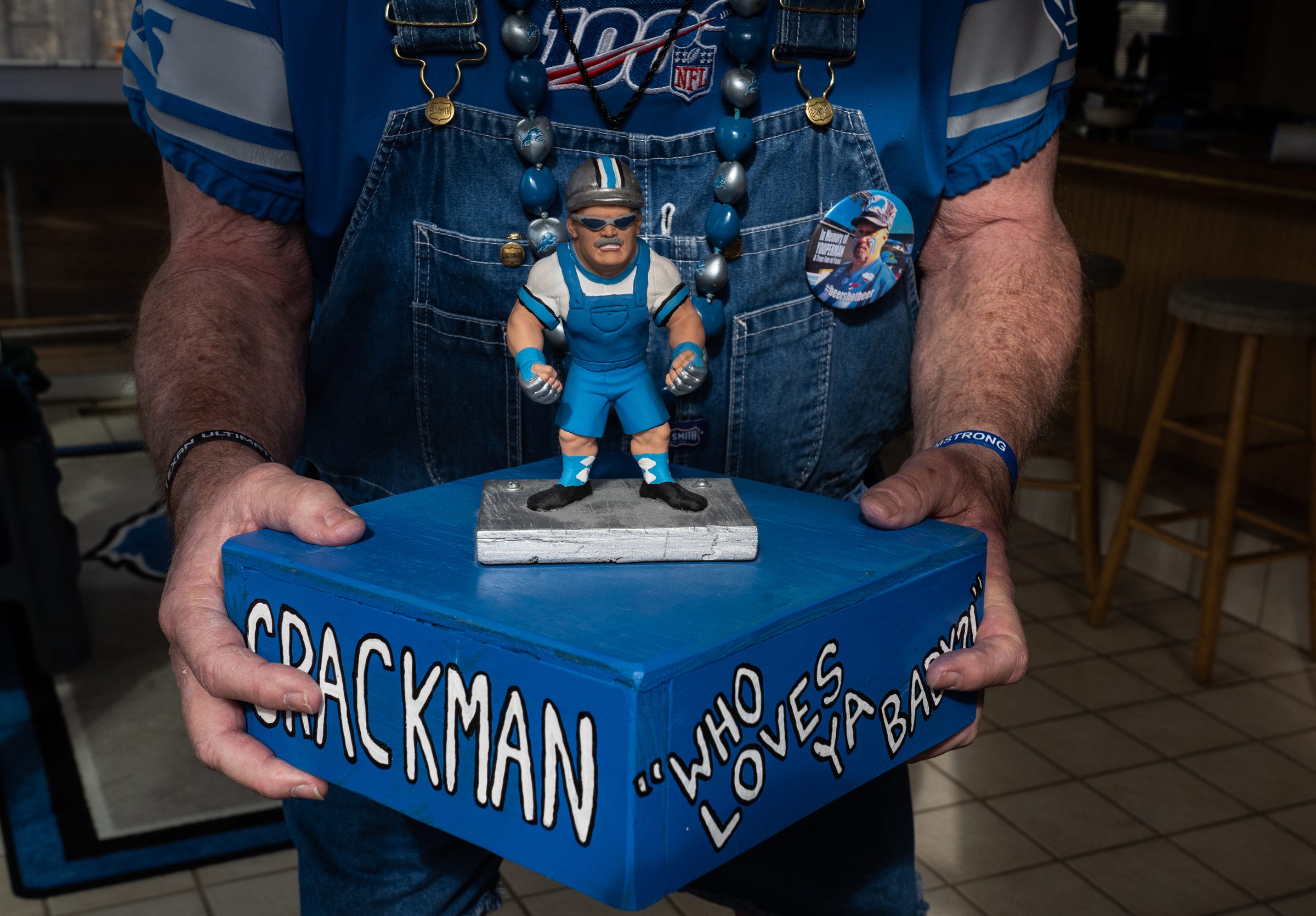 Diehard Detroit Lions Superfan Ron "Crackman" Crachiola, 69, of Macomb Township, holds a figurine a friend made for him with his slogan "Who loves ya, baby?" While wearing a button in memory of his friend and Detroit Lions fan the late Donnie "Yooperman" Stefanski at his home in Macomb Township on March 3, 2021.