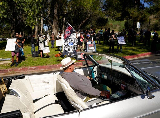 FILE - In this Feb. 27, 2021, file photo, a driver in his convertible cruises past a small group of anti-COVID-19 vaccine protesters demonstrating at Elysian Park, outside the Dodger Stadium vaccination mass center in Los Angeles. As the world struggles to break the grip of COVID-19, psychologists and misinformation experts are studying why the pandemic spawned so many conspiracy theories, which have led people to eschew masks, social distancing and vaccines. (AP Photo/Damian Dovarganes, File)