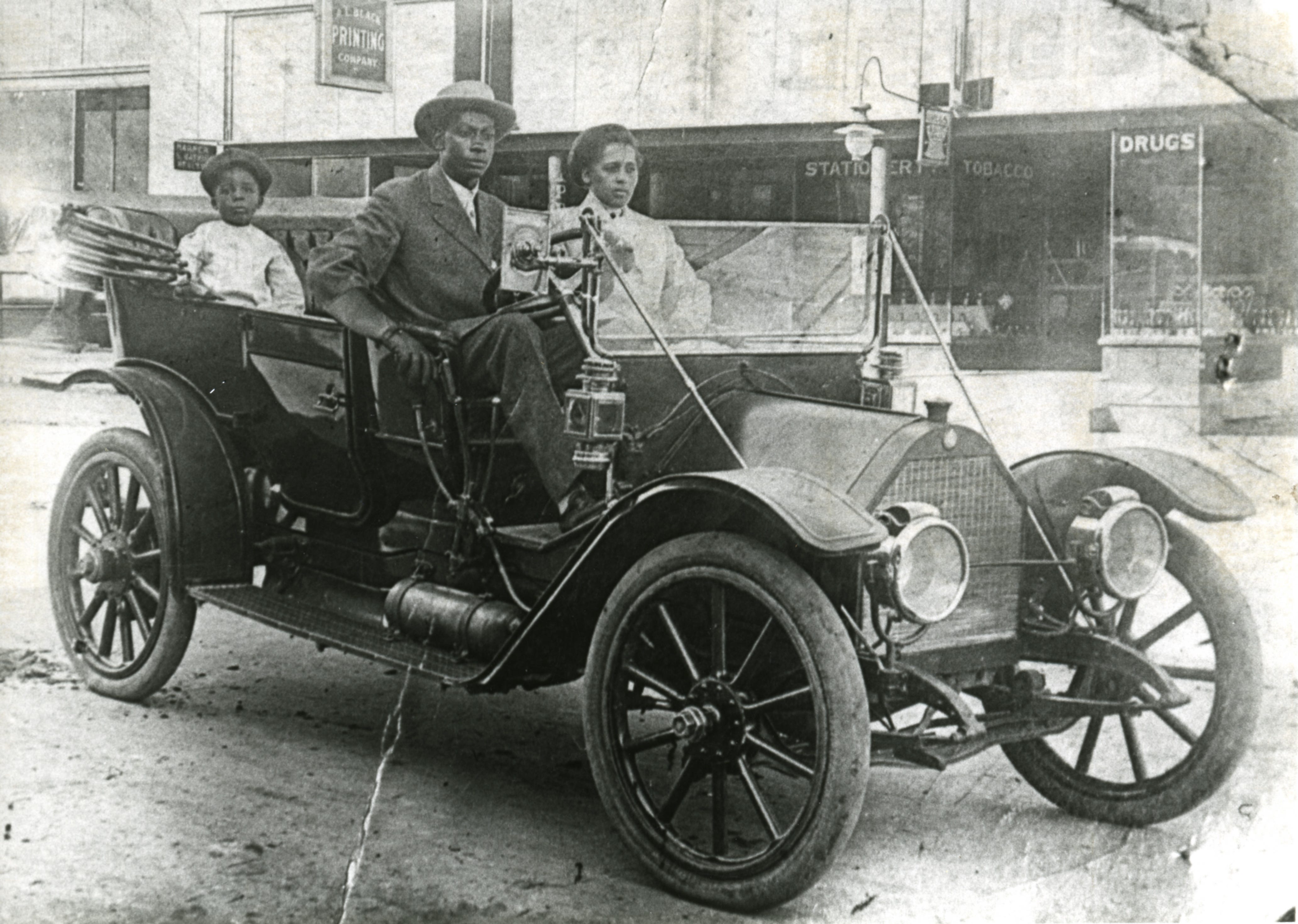 John Wesley Williams and wife, Loula Cotten Williams, and their son, William Danforth Williams, sitting in a 1911 Norwalk automobile. A sign advertising A.L. Black Printing Co., located at 114 S Boston Ave., Tulsa, is visible in the background. John was an engineer for Thompson Ice Cream Co. Loula was a teacher in Fisher. The Williams family owned the Dreamland Theatre, which opened in 1914 at 129 N Greenwood Ave., and was destroyed in the 1921 Tulsa Race Massacre.