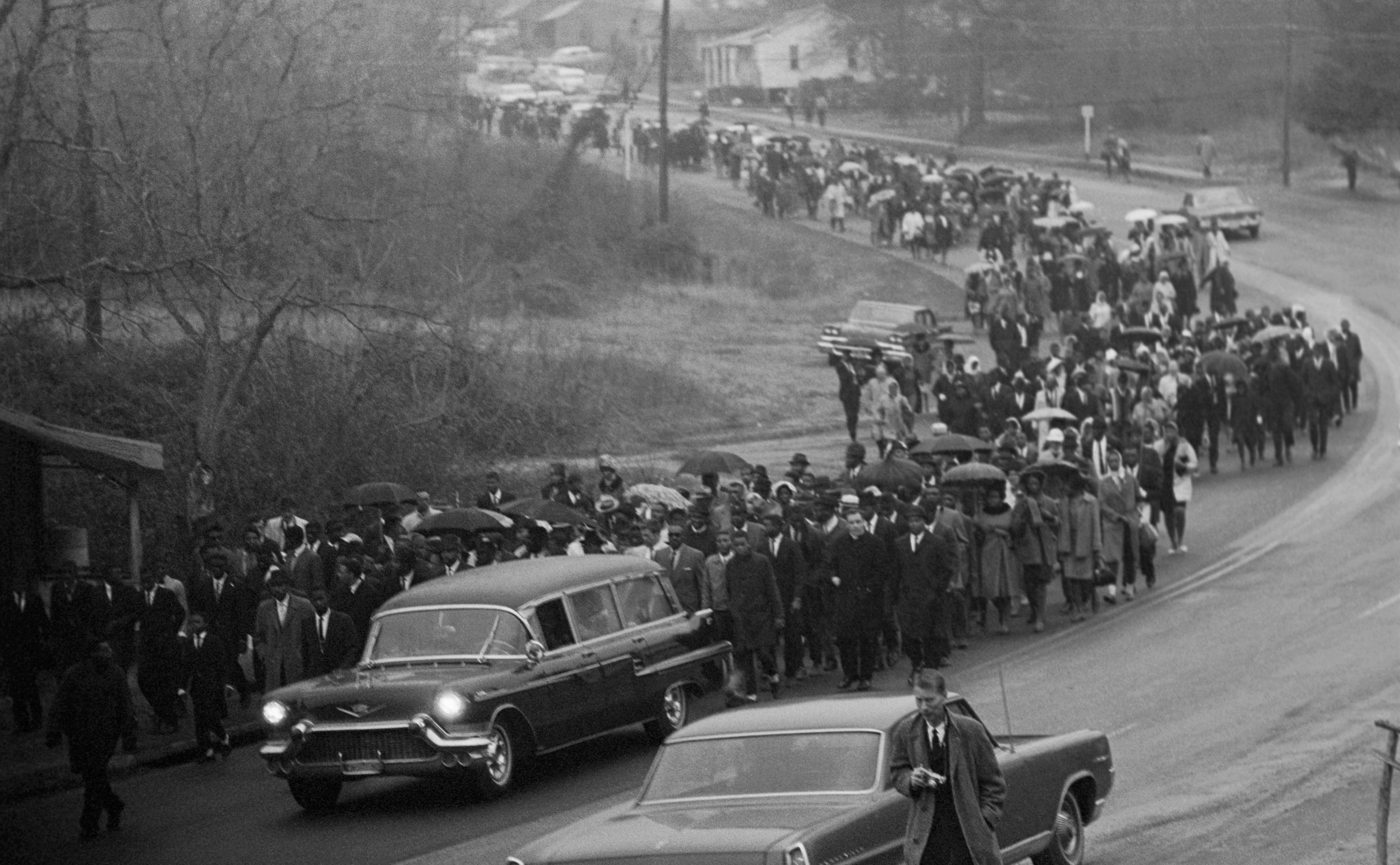 A hearse carrying the body of Jimmie Lee Jackson drives slowly in rain with an estimated 700 people, mostly African American, following the cemetery where Jackson is to be lain. Jackson was shot in Marion February 18, during a night demonstration and died in Selma Hospital. Two funerals were held for the civil rights protester with Dr. Martin Luther King giving the eulogy.