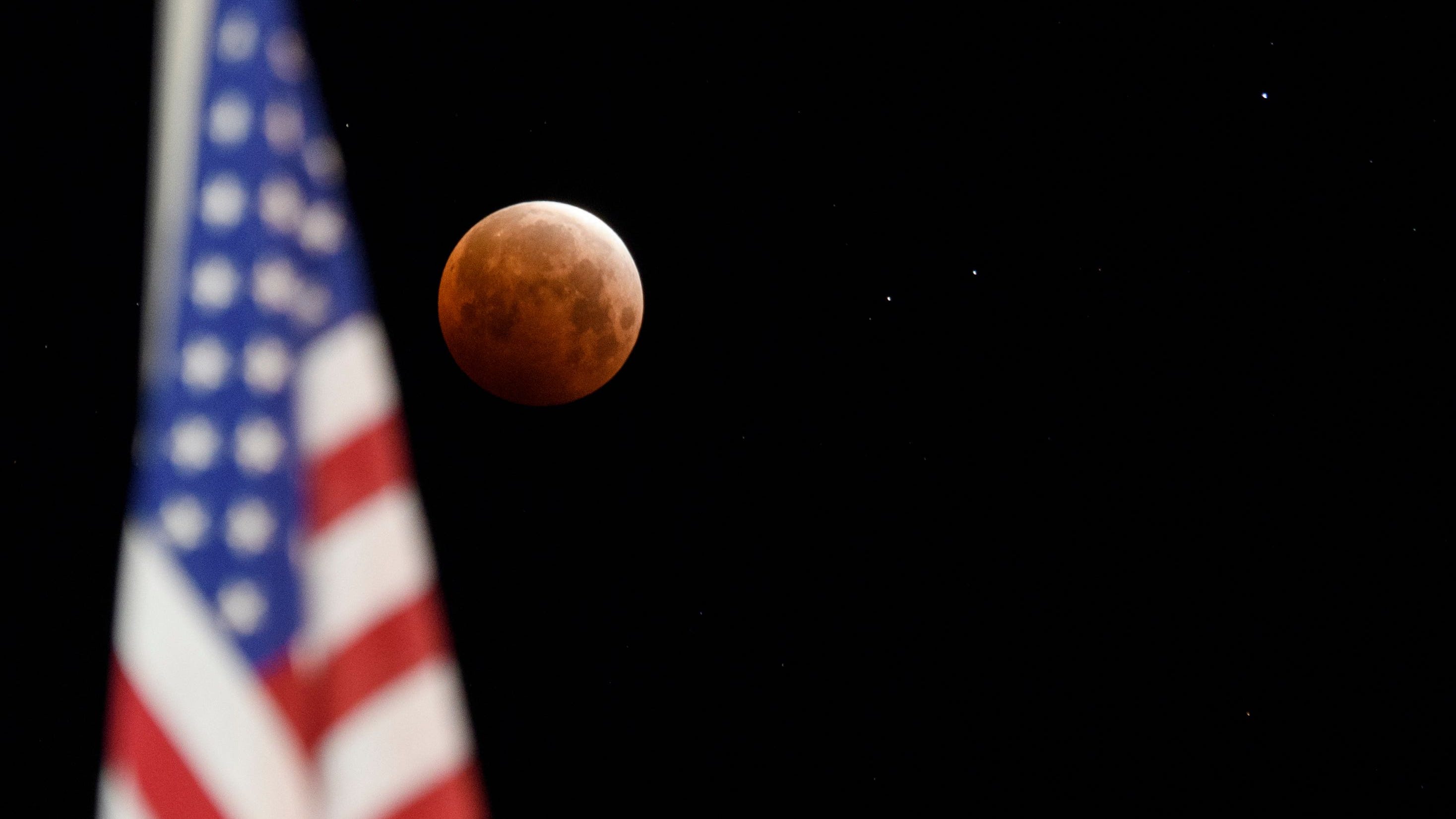 Una luna llena se acerca a la totalidad enmarcada por una bandera de los EE. UU. Durante un eclipse lunar total cuando la luna se adentra en la sombra de la tierra para una superluna de sangre el 26 de mayo de 2021 en Chico, California.