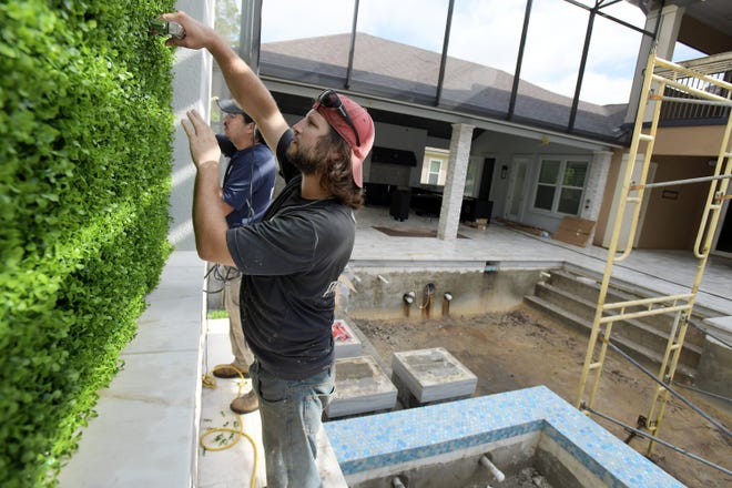 Jamie Cuneo, a maker for the Pratt Guys, and crew leader Mark Ruel Jr. install an artificial boxwood wall covering behind a hot tub on a home-renovation project in the Deerwood neighborhood of Jacksonville in June. The project includes a pool with a hot tub and sunken fire pit, a large covered outdoor kitchen, marble tile and a basketball court, all covered by a towering screened-in enclosure.