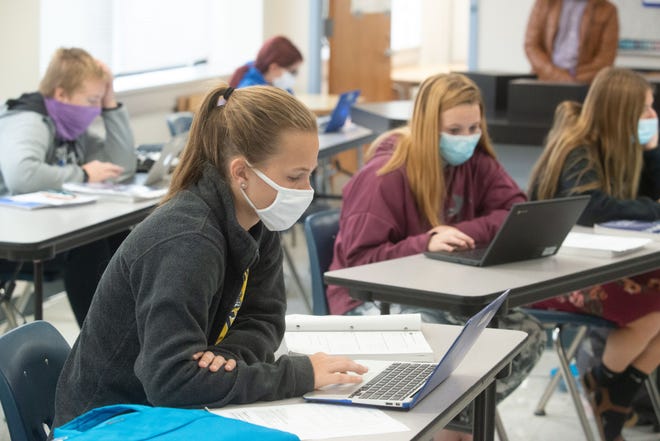 Students wearing masks working on laptops at classroom tables.