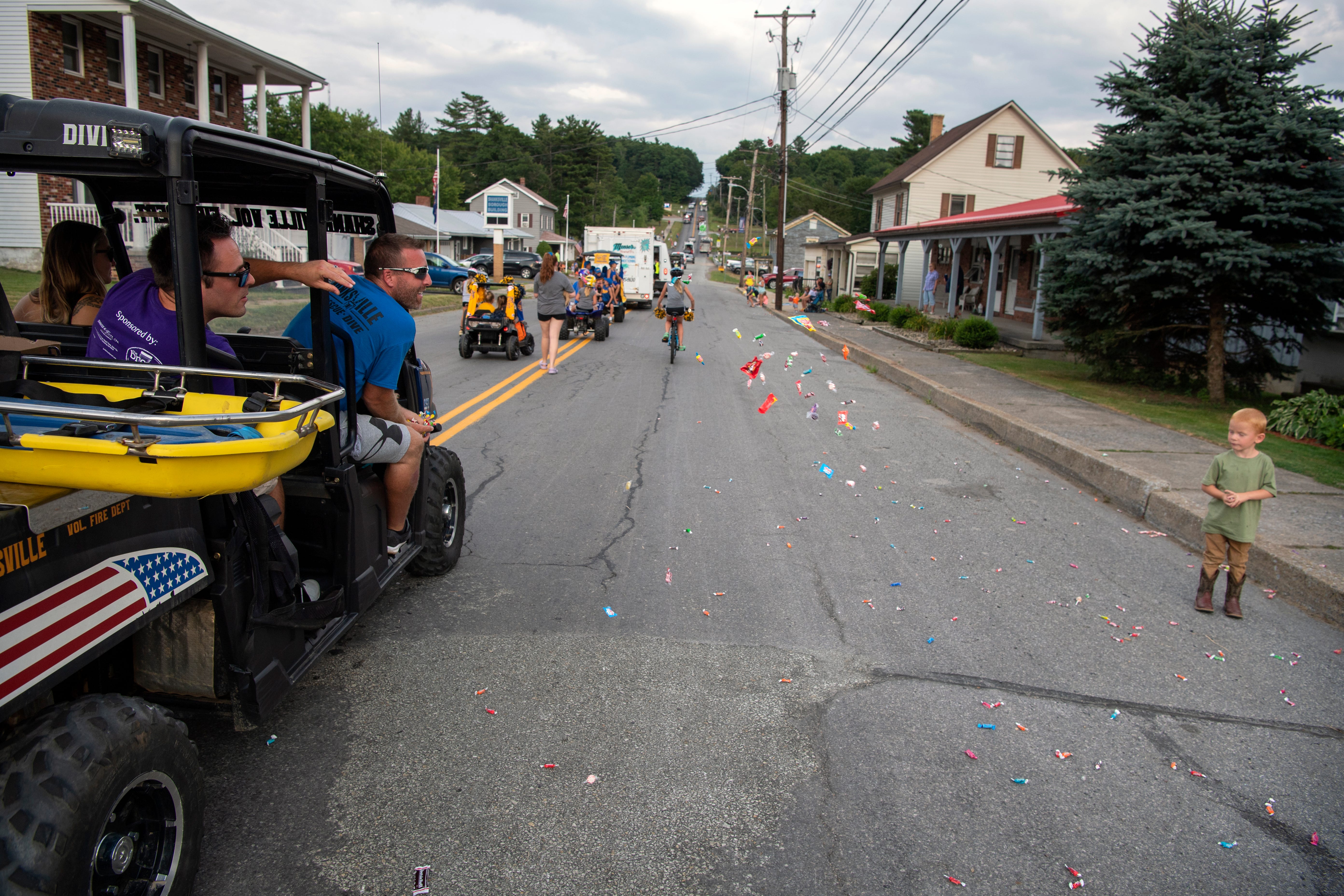 (Left) Shanksvillel Mayor Chris Baeckel throws candy to Boyd Benning, 3, during the Volunteer Fire Department parade on Main Street in Shanksville, PA, on Friday, August 6, 2021. (Second from left) John Abramowich sits in front of Baeckel. 