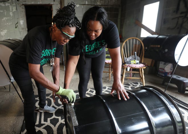 Deborah Turner, 63 and her daughter, Larenda Walker, 34 show how they use chalk to mark guide lines mark the door of the grill on steel drums barrel that will  turned into bbq grill in the garage of her home on Thursday, Aug. 12, 2021, in Akron. The mother and daughter duo are carrying on the tradition of making the grills in honor of her father George Hammett, 93 who died early this year of COVID.