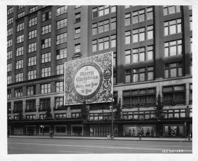 Black and white photographic print, taken from the southwest side of Woodward Avenue, and looking toward the Woodward Avenue entrance of The J.L. Hudson Company Department Store in 1941. A large Christmas sign is affixed to the exterior of the department store and features holiday ornamentation; the sign reads, "A Merry Christmas and a Happy New Year to You."