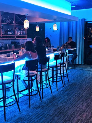The bar at Kyle G's Oyster & Wine Bar is faced with white subway tile and light blue under counter lighting with gold speckled pendants and simple cushioned stools.