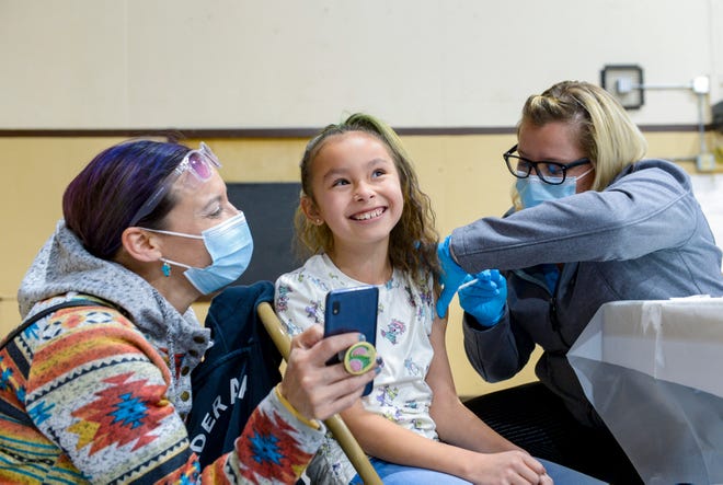 Ivoree Morsette, age 7, is all smiles while receiving her COVID vaccine from nurse Amanda Dement while her mother Billi Raining Bird attempts to distract her with funny messages on her phone, Wednesday evening during Alluvion Health's vaccine clinic at Paris Gibson Education Center.