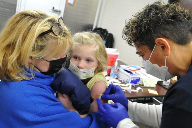 Brewster Ambulance licensed practical nurse Tara Mendonca, right, places a bandage on the arm of Peggy Lyons, 5, of Weymouth, center, as she is comforted by her mom, Cindy Lyons, left, during the COVID-19 student vaccination clinic at Weymouth High School on Saturday, Nov. 20, 2021.