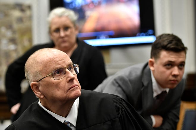 Judge Bruce Schroeder, front,  comes down from the bench and sits closer to a 4k television screen to watch a video as defendant Kyle Rittenhouse, right, and his attorney Natalie Wisco stand behind him during proceedings at the Kenosha County Courthouse on Nov. 12, 2021, in Kenosha, Wisconsin. (Sean Krajacic/Pool/Getty Images/TNS)