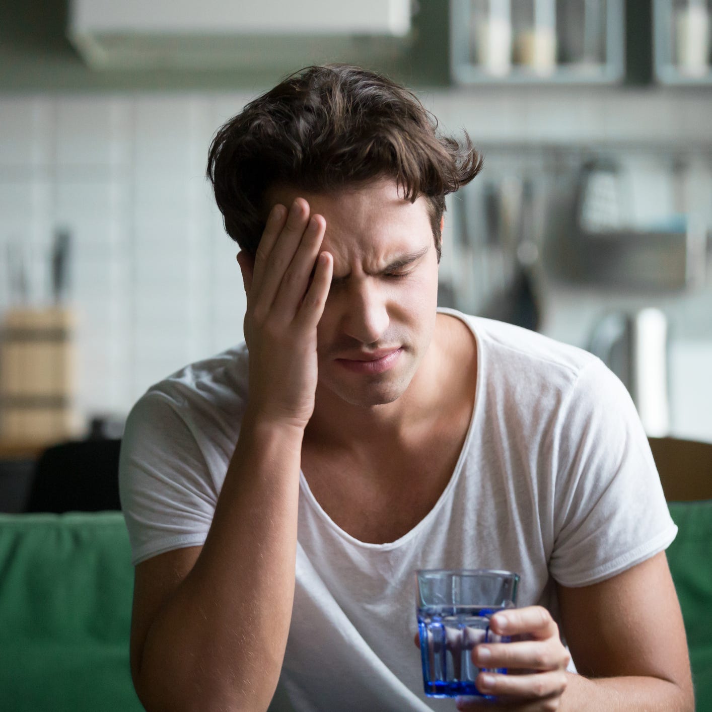 Young man suffering from strong headache, sitting with glass of water in the kitchen,