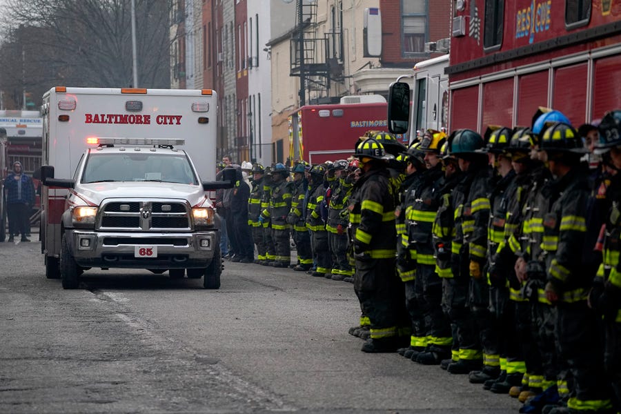Firefighters salute as an ambulance carries a deceased firefighter after they were pulled out of a collapsed building while battling a two-alarm fire at a vacant row home, Monday, Jan. 24, 2022, in Baltimore. Officials said several firefighters died during the blaze. (AP Photo/Julio Cortez)