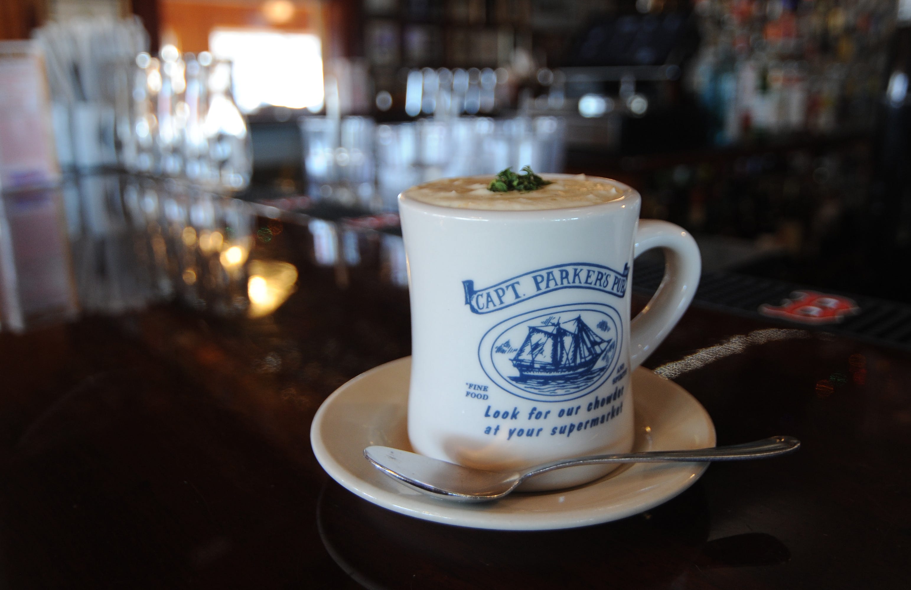 A white mug that has the Captain Parker logo on it filled to overflowing with New England clam chowder in the foreground with an out-of-focus bar in the background