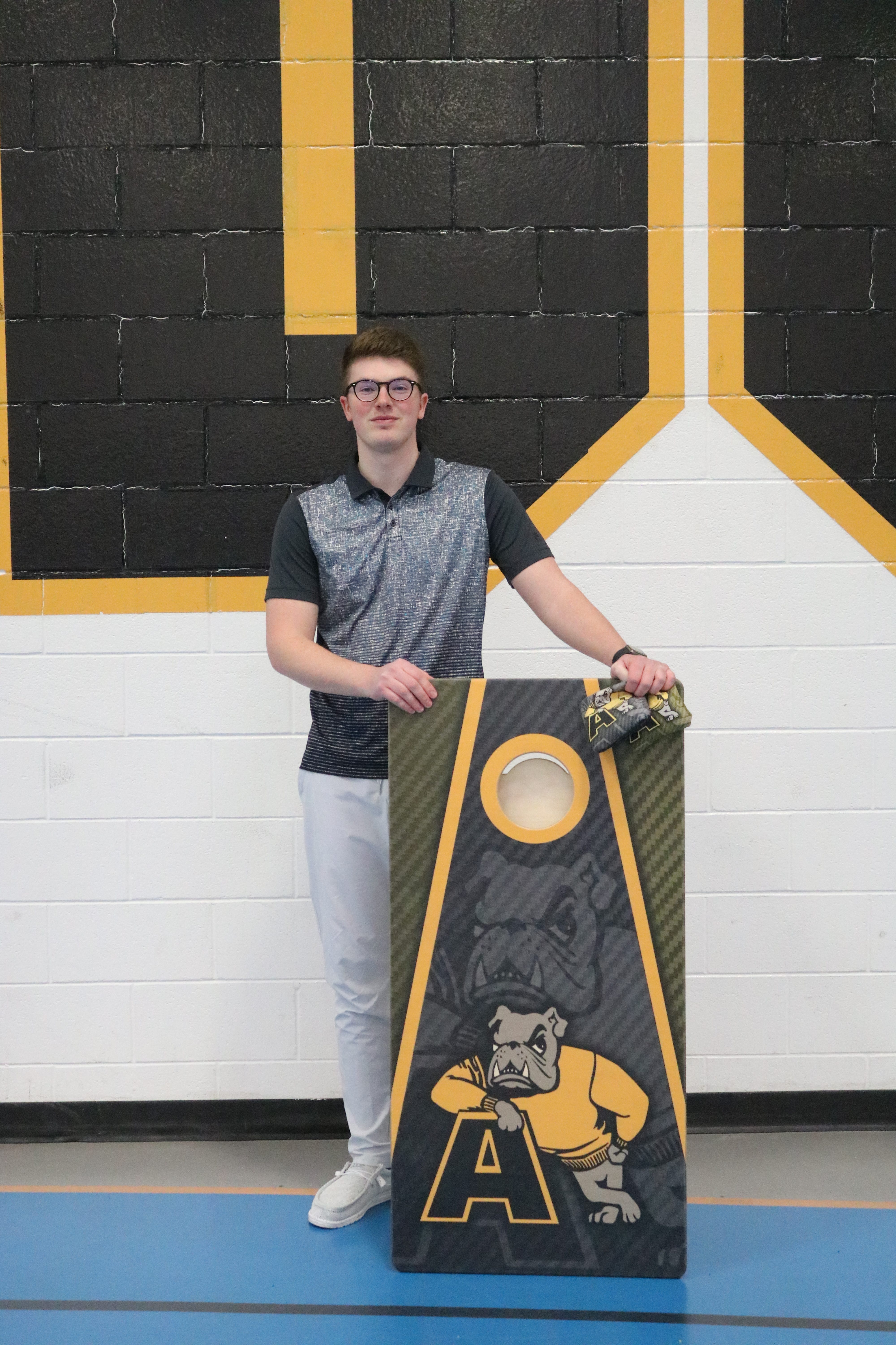 Adrian College cornhole coach Max Benedict (Tecumseh) stands with a pair of cornhole bags and a cornhole board emblazoned with the Adrian colors on Monday, Feb. 14, at the Merillat Sport and Fitness Center.