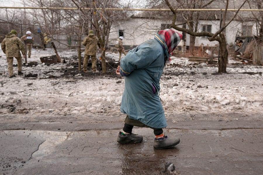 An elderly lady walks by as members of the Joint Centre for Control and Coordination on ceasefire of the demarcation line, or JCCC, survey a crater and damage to a house from artillery shell that landed in Vrubivka, Ukraine, one of the at least eight that hit the village today, according to local officials, in the Luhansk region, eastern Ukraine, Thursday, Feb. 17, 2022. U.S. President Joe Biden warned that Russia could still invade Ukraine within days and Russia expelled the No. 2 diplomat at   the U.S. Embassy in Moscow, as tensions flared anew in the worst East-West standoff in decades.