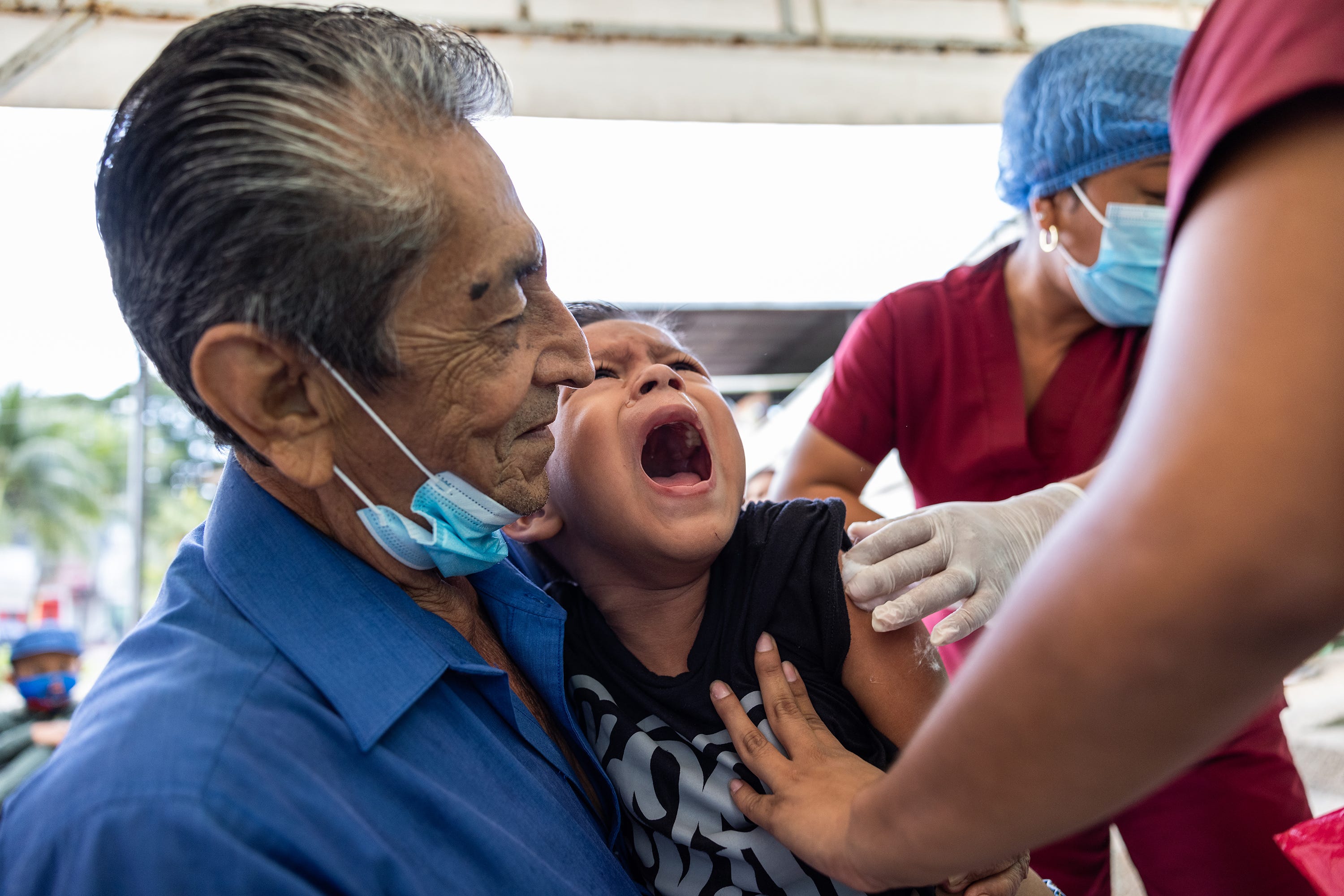 A child screams in fear at the sight of the syringes. The nurses, with the help of his grandfather, try to calm him down in order to proceed with the vaccination.