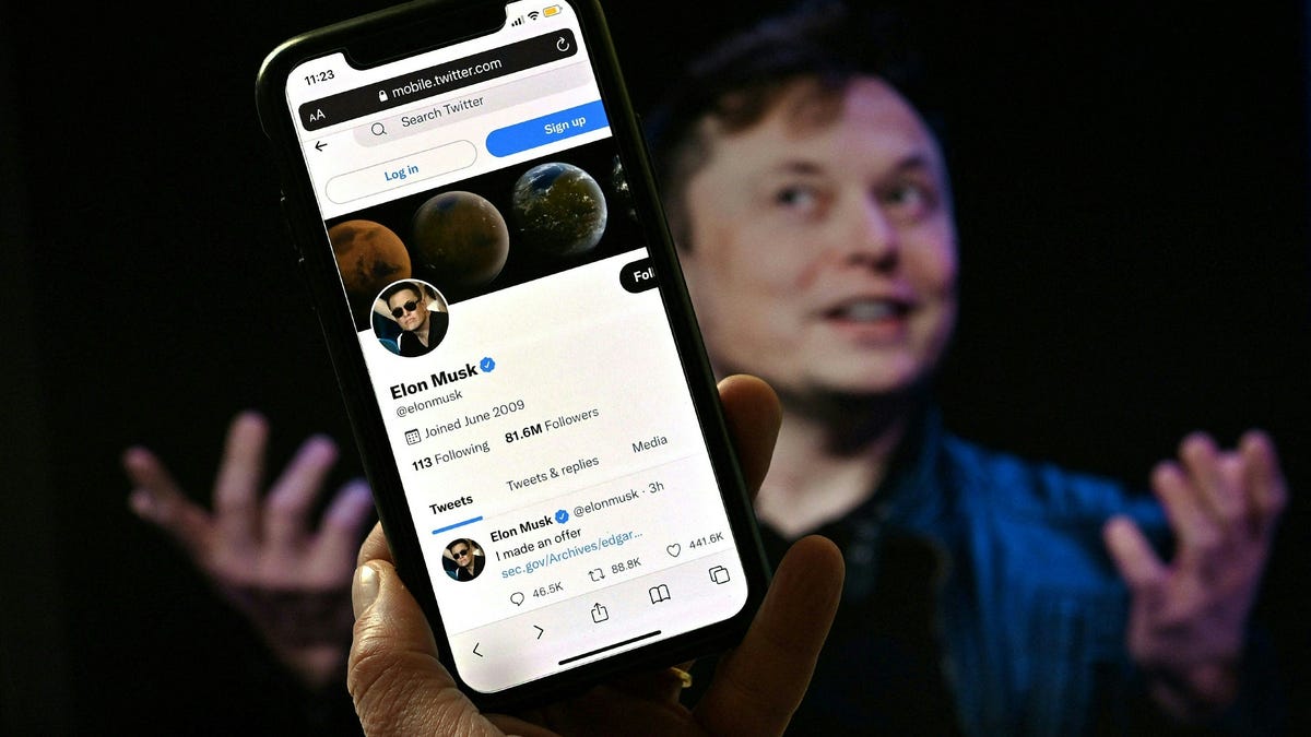 A phone screen displays the Twitter account of Elon Musk with a photo of him shown in the background