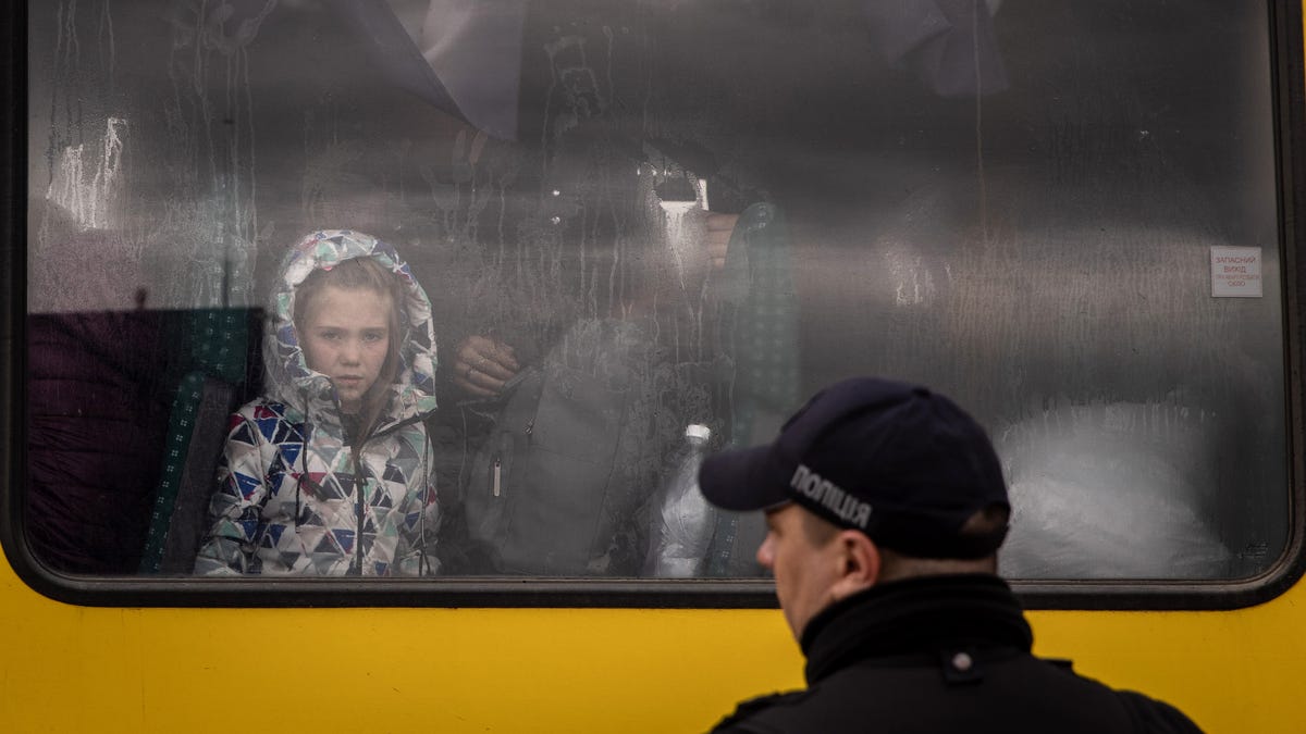A police officer looks on as a girl from Mariupol looks out the window of a bus after a convoy of vehicles arrived at an evacuation point, carrying people from Mariupol, Melitopol and surrounding towns under Russian control on April 21, 2022 in Zaporizhzhia, Ukraine.