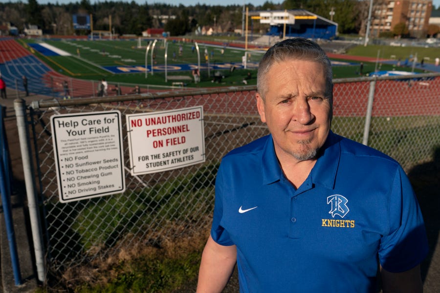 Joe Kennedy, a former assistant football coach at Bremerton High School in Bremerton, Wash., poses for a photo March 9, 2022, at the school's football field. After losing his coaching job for refusing to stop kneeling in prayer with players and spectators on the field immediately after football games, Kennedy will take his arguments before the U.S. Supreme Court on Monday, April 25, 2022, saying the Bremerton School District violated his First Amendment rights by refusing to let him continue   praying at midfield after games. Kennedy said signs like the one on the fence behind him were put up after he started praying after games with players in efforts to keep spectators from rushing the field to join them. (AP Photo/Ted S. Warren) ORG XMIT: WATW303