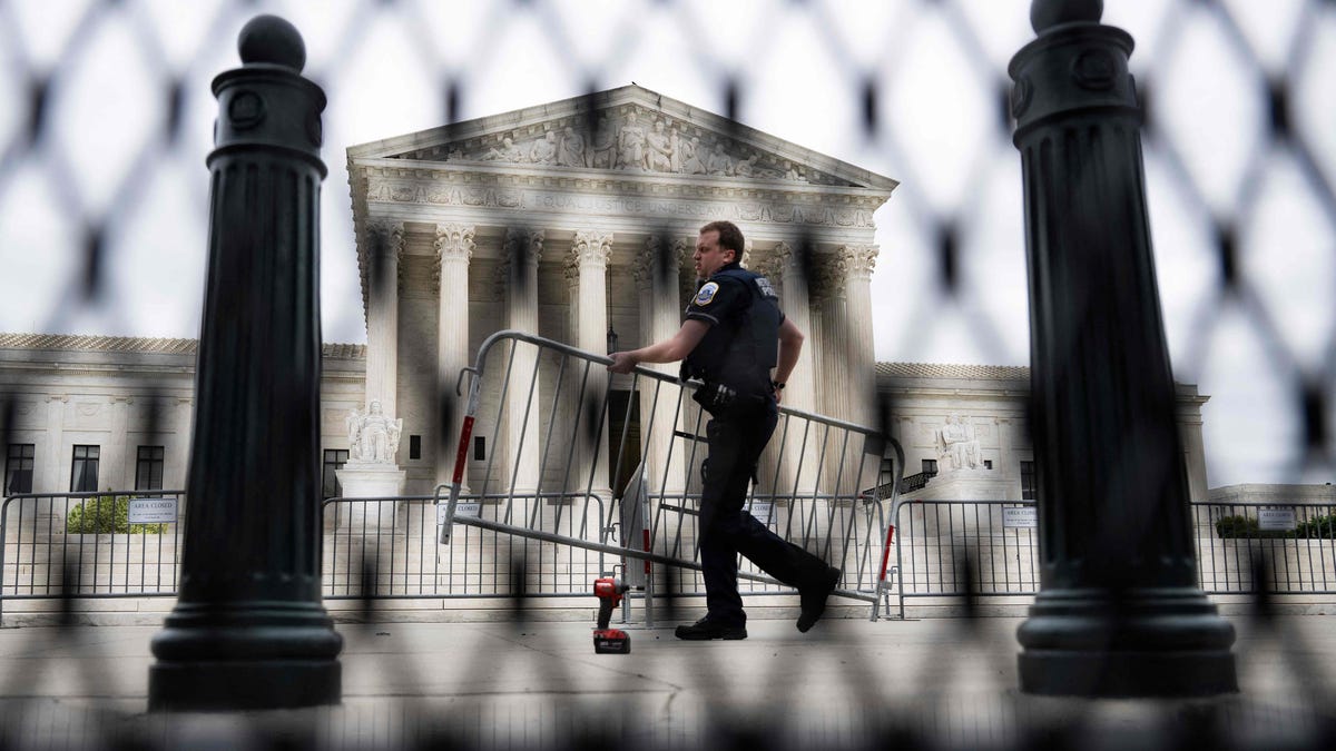 A police officer carrying a barricade is seen through un-scalable fence that stands around the US Supreme Court in Washington, DC, on May 5, 2022. - The fencing is being set up due to the protests that have occurred in response to the leaked draft of a majority opinion that would shred nearly 50 years of constitutional protections. The draft, obtained by Politico, was written by Justice Samuel Alito, and has been circulated inside the conservative-dominated court,   the news outlet reported. Politico stressed that the document it obtained is a draft and opinions could change. The court is expected to issue a decision by June (Photo by Jim WATSON / AFP) (Photo by JIM WATSON/AFP via Getty Images) ORG XMIT: 0 ORIG FILE ID: AFP_329J687.jpg
