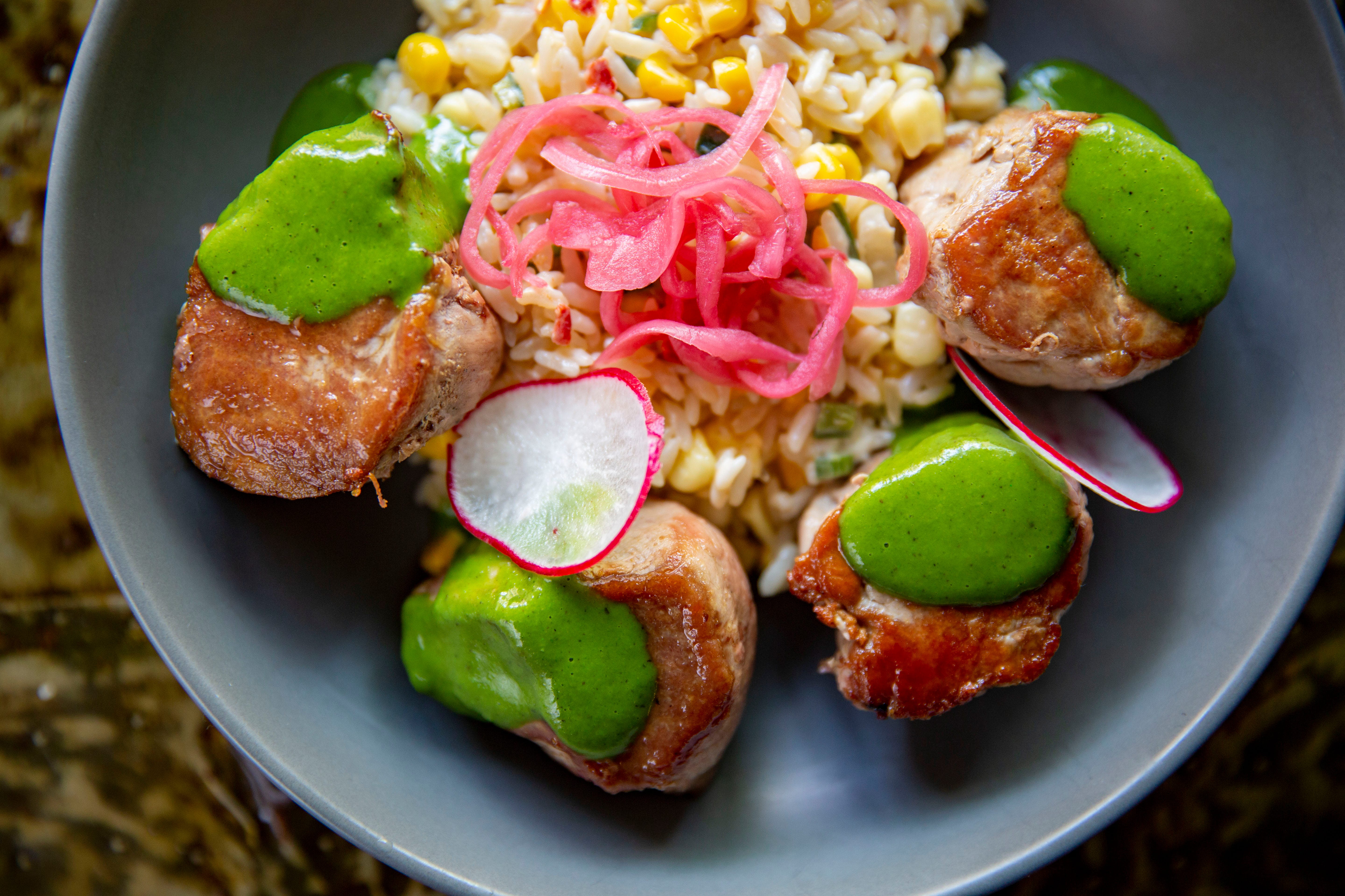 Four pork medallions with green sauce and thinly sliced radishes on top. In the center, brown rice with pink marinated onions on top. 