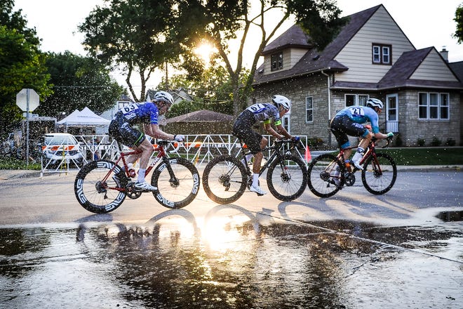 A group of riders passes a sprinkler set up at the corner of S. 68th Street and W. Rogers Street during the West Allis Cheese Wheel Classic  June 21, 2022, in West Allis. The temperature at the start of the race was nearly 100 degrees.